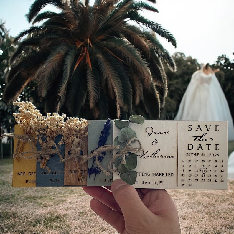 May include: A set of wooden save the date cards with dried flowers and greenery. The cards are tied together with twine and have the text "James & Katherine" and "Save the Date June 11, 2025" on them.
