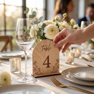 May include: An arched wooden table number sign featuring the text Table 4 in a black script and serif font, decorated with delicate line art of floral branches on both sides, standing upright on a rectangular wooden base on a dining table.