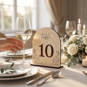 May include: An arched wooden table number sign featuring the text Table 10 in a dark script and block font, accented with delicate black line-drawn floral sprigs on the sides, standing on a rectangular base on a set dining table.