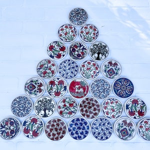 May include: A triangular arrangement of 21 round ceramic coasters with intricate floral and geometric designs in shades of blue, red, and white. The coasters are arranged in a pyramid shape on a white brick wall.