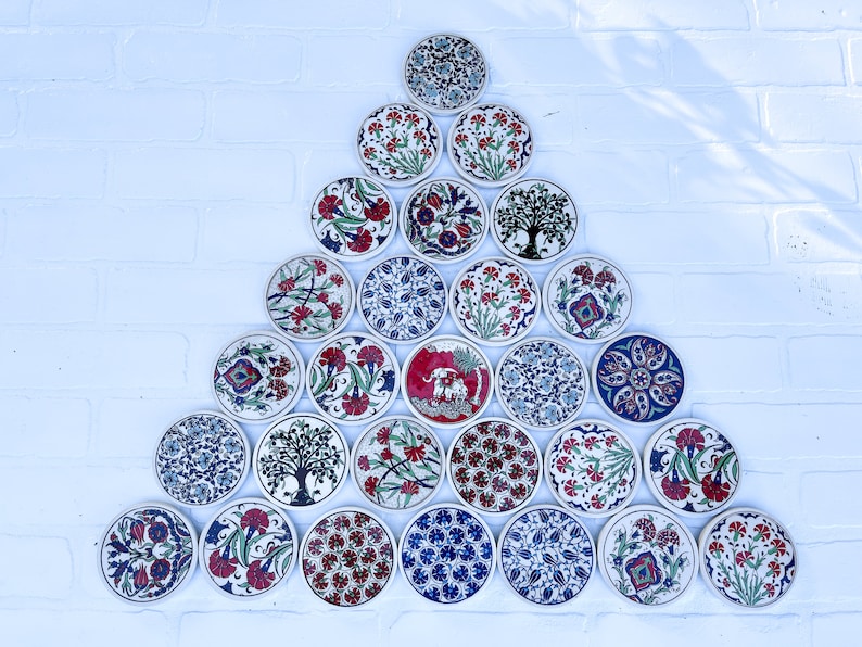 May include: A triangular arrangement of 21 round ceramic coasters with intricate floral and geometric designs in shades of blue, red, and white. The coasters are arranged in a pyramid shape on a white brick wall.