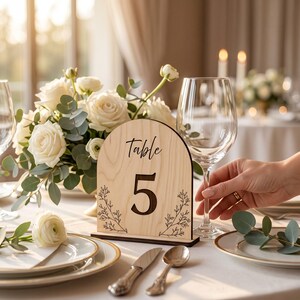 May include: An arched light wood table number sign featuring the text Table 5 in a black script and print font, decorated with black line art of botanical sprigs on both sides, standing on a dark wood base on a white tablecloth.