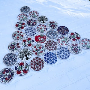 May include: A collection of 24 round ceramic coasters with intricate floral and geometric designs in shades of blue, red, and white. The coasters are arranged in a triangular pattern on a white surface.