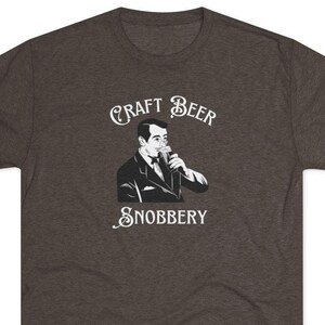 Può includere: Una t-shirt color grigio scuro melange con una grafica bianca di un uomo che beve birra. Il testo "CRAFT BEER SNOBBERY" è stampato sulla t-shirt.