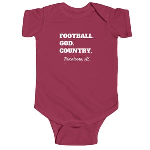Puede incluir: Un body de bebé de color rojo carmesí con texto blanco que dice "FOOTBALL. GOD. COUNTRY. Tuscaloosa, AL".