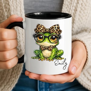 May include: White ceramic mug with a black interior and handle, featuring a cartoon frog wearing glasses, a bow tie, and a leopard print bow. The name "Emily" is written in cursive on the mug.