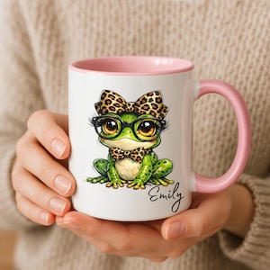 A close-up of hands holding a white ceramic mug featuring a light pink handle and inner rim accent. The surface displays a personalized funny frog mug design with a cute whimsical frog illustration complete with big eyes, black-rimmed glasses, a leopard print hair bow, and a matching leopard print bowtie. The custom name "Emily" is printed in an elegant black cursive script below. highlighting this quirky animal ceramic mug as a perfect custom gift for her.