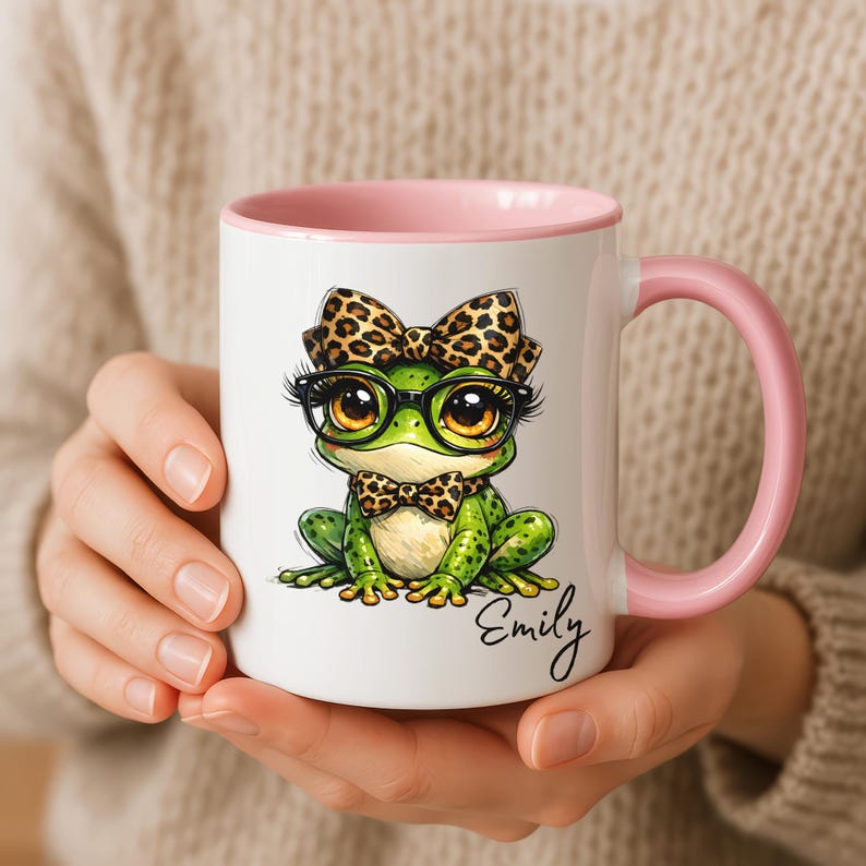 A close-up of hands holding a white ceramic mug featuring a light pink handle and inner rim accent. The surface displays a personalized funny frog mug design with a cute whimsical frog illustration complete with big eyes, black-rimmed glasses, a leopard print hair bow, and a matching leopard print bowtie. The custom name "Emily" is printed in an elegant black cursive script below. highlighting this quirky animal ceramic mug as a perfect custom gift for her.