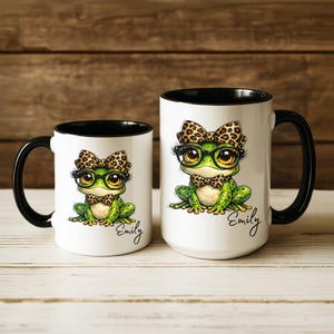 Puede incluir: Dos tazas de cer&aacute;mica blanca con interiores y asas negras. Cada taza presenta una rana de dibujos animados con gafas, pajarita y lazo, todo con un dise&ntilde;o de estampado de leopardo. El nombre "Emily" est&aacute; escrito en cursiva en cada taza.