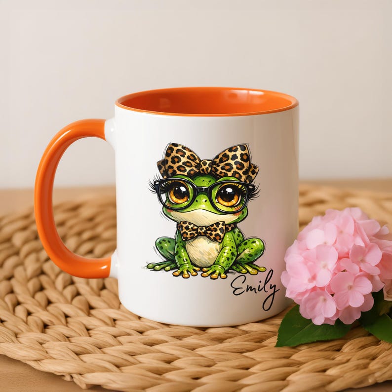 Puede incluir: Taza de cer&aacute;mica blanca con interior y asa naranja. La taza presenta una rana de dibujos animados con gafas y pajarita, ambas con dise&ntilde;o de estampado de leopardo. La rana es verde con ojos grandes. El nombre "Emily" est&aacute; escrito en la taza.