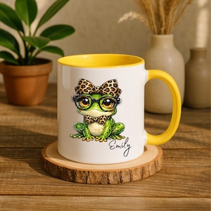 A white ceramic mug featuring a bright yellow handle and inner rim accent sits on a natural wood slice coaster. The mug displays a personalized funny frog mug design with a cute whimsical frog illustration wearing large glasses, a leopard print bow, and a matching bowtie. The custom name "Emily" is cleanly written in black cursive script below the animal artwork. The setting is a rustic wooden table styled with a plant and a ceramic vase, making this coffee cup an excellent custom gift for her.