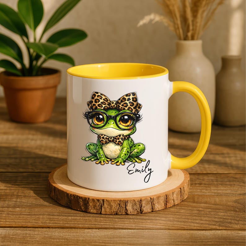 A white ceramic mug featuring a bright yellow handle and inner rim accent sits on a natural wood slice coaster. The mug displays a personalized funny frog mug design with a cute whimsical frog illustration wearing large glasses, a leopard print bow, and a matching bowtie. The custom name "Emily" is cleanly written in black cursive script below the animal artwork. The setting is a rustic wooden table styled with a plant and a ceramic vase, making this coffee cup an excellent custom gift for her.