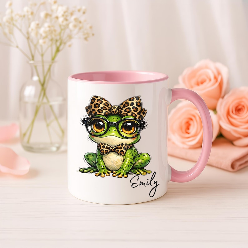 A white ceramic mug with a light pink handle and inner rim stands on a tabletop surrounded by romantic pink roses and delicate flowers in a small glass vase. The mug features a personalized funny frog mug design with a cute whimsical frog illustration sporting big eyes, black glasses, a leopard print bow, and a matching bowtie. The custom name "Emily" is cleanly written in black cursive script below the animal print artwork, highlighting this coffee cup as a perfect personalized gift for her.