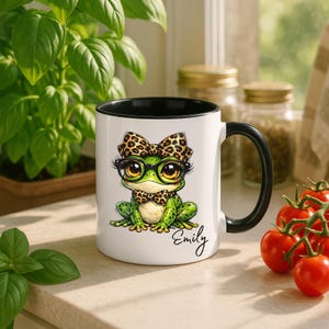 A white ceramic mug with a black handle and inner rim stands on a, rustic wood slice coaster. The mug features a personalized funny frog mug design with a cute whimsical frog illustration wearing large glasses, a leopard print bow, and a matching bowtie. The custom name "Emily" is printed in a black cursive script below the animal artwork. this coffee cup as an ideal personalized gift for her or any frog lover.