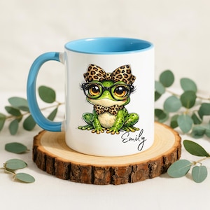 A white ceramic mug featuring a vibrant light blue handle and inner rim accent sits on a natural wood slice coaster surrounded by eucalyptus leaves. The mug showcases a personalized funny frog mug design with a cute whimsical frog illustration wearing black glasses, a leopard print hair bow, and a matching leopard print bowtie. The custom name "Emily" is neatly printed in black cursive script below the animal artwork, making this quirky ceramic mug a lovely personalized gift for her.