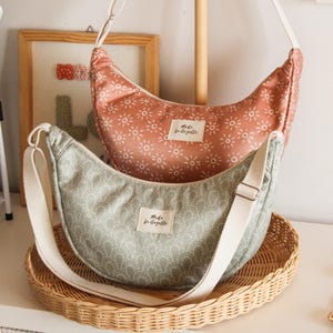 Può includere: Due borse a tracolla a forma di mezzaluna. Una è color rosa antico con un motivo a raggiera bianco. L'altra è verde salvia con un motivo a ventaglio bianco. Entrambe le borse hanno una tracolla bianca regolabile e una piccola etichetta "Moda la Gazelle".