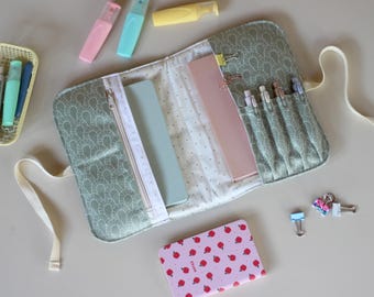 May include: An open, sage green fabric pencil case with a white zipper, holding notebooks and pens. The case is surrounded by highlighters, a small notebook with a strawberry pattern, and binder clips.