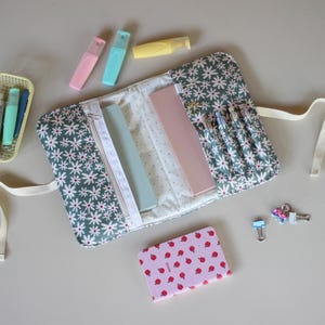 May include: A daisy-patterned pencil case with a zipper, holding pens and rulers. Pastel highlighters and a small notebook with a strawberry design are nearby. A small basket with pens and clips are also visible.