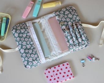 May include: A daisy-patterned pencil case with a zipper, holding pens and rulers. Pastel highlighters and a small notebook with a strawberry design are nearby. A small basket with pens and clips are also visible.