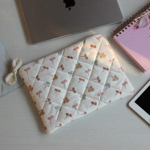 May include: A quilted laptop sleeve with a floral pattern in pastel colors. The sleeve is white with a grid pattern and features small flowers in pink, orange, and blue. A laptop, notebook, and tablet are also visible.