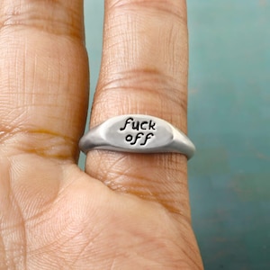 May include: A silver ring with the words "fuck off" engraved on the oval face. The ring is worn on a finger, and the text is in a handwritten style.