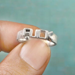 May include: A silver ring with the word "FUCK" spelled out in block letters. The ring is held between two fingers against a teal background. The ring is made of a shiny metal.