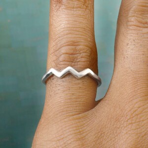 May include: A silver ring featuring a zigzag pattern. The ring, worn on a finger, has a contemporary design. The ring's design comprises connected chevrons, forming a wave-like shape. The ring is crafted from a polished, reflective metal, showcasing a minimalist style.
