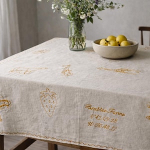 Op de afbeelding: Een beige linnen tafelkleed met gouden borduursels van fruit, groenten en tekst. Een kom met citroenen en een vaas met bloemen staan op de tafel. Het tafelkleed heeft een geschulpte rand.