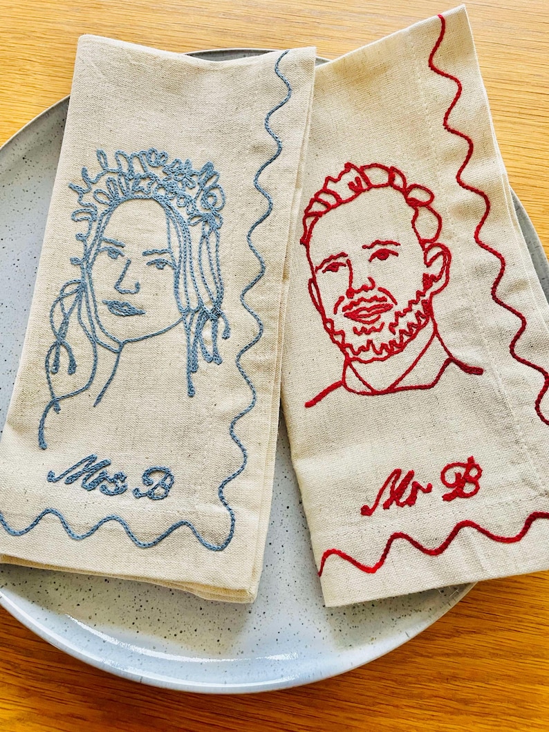 Puede incluir: Dos servilletas de lino con retratos bordados. Una servilleta presenta el rostro de una mujer bordado en azul con una corona floral y el texto "Mrs B". La otra servilleta tiene el rostro de un hombre bordado en rojo y el texto "Mr B". Ambas tienen un borde ondulado.