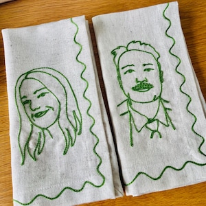 Puede incluir: Dos servilletas de lino blanquecino con retratos bordados en verde de una mujer y un hombre. Los retratos est&aacute;n delineados con hilo verde, con un borde verde ondulado. Las servilletas est&aacute;n sobre una superficie de madera.