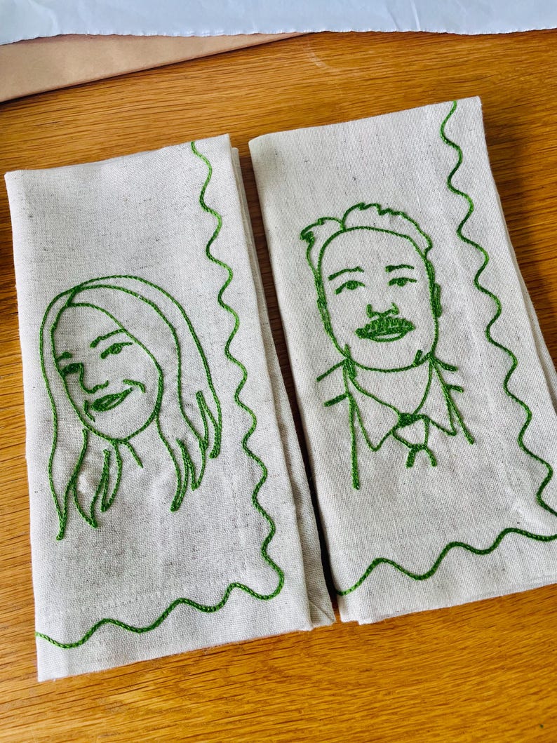 Puede incluir: Dos servilletas de lino blanquecino con retratos bordados en verde de una mujer y un hombre. Los retratos est&aacute;n delineados con hilo verde, con un borde verde ondulado. Las servilletas est&aacute;n sobre una superficie de madera.