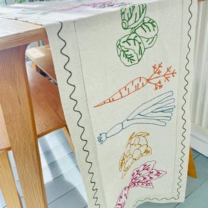 May include: Cream-coloured table runner with embroidered vegetables. Features green cabbage, orange carrot, blue leek, yellow artichoke, and red beet. The edges are trimmed with a black zigzag stitch. The runner is draped over a wooden table.