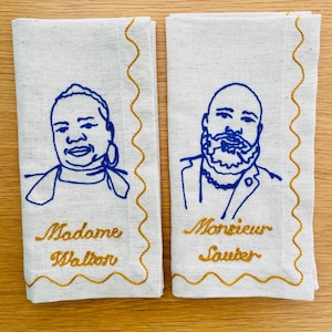 Puede incluir: Dos servilletas de lino blanquecino con retratos bordados en azul y bordes festoneados dorados. Una servilleta dice "Madame Walton" y la otra "Monsieur Sauter" en letras doradas. Las servilletas est&aacute;n sobre una superficie de madera clara.