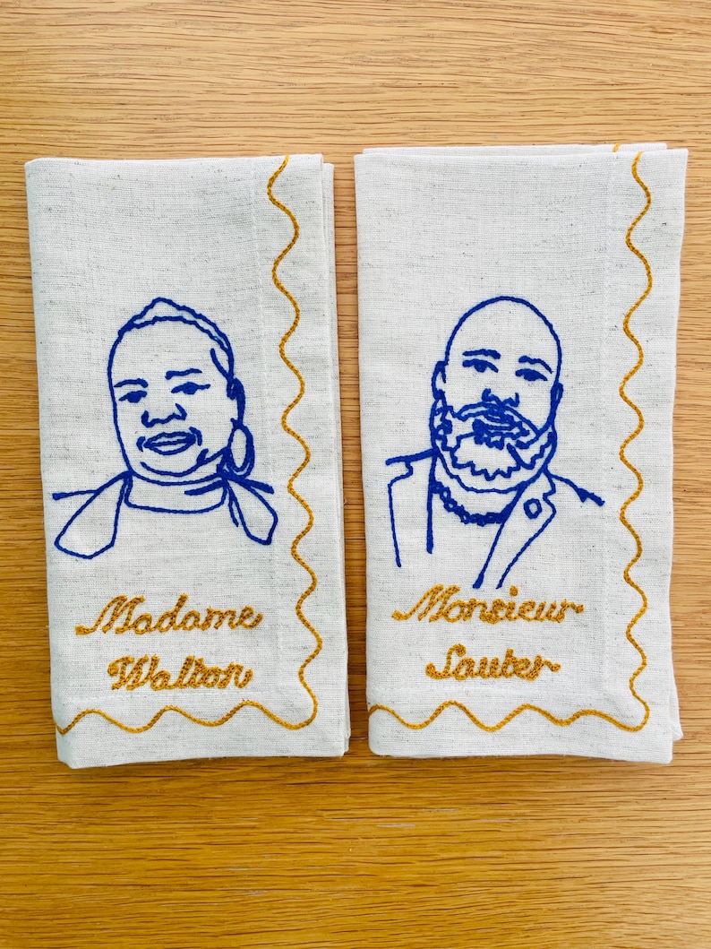 Puede incluir: Dos servilletas de lino blanquecino con retratos bordados en azul y bordes festoneados dorados. Una servilleta dice "Madame Walton" y la otra "Monsieur Sauter" en letras doradas. Las servilletas est&aacute;n sobre una superficie de madera clara.