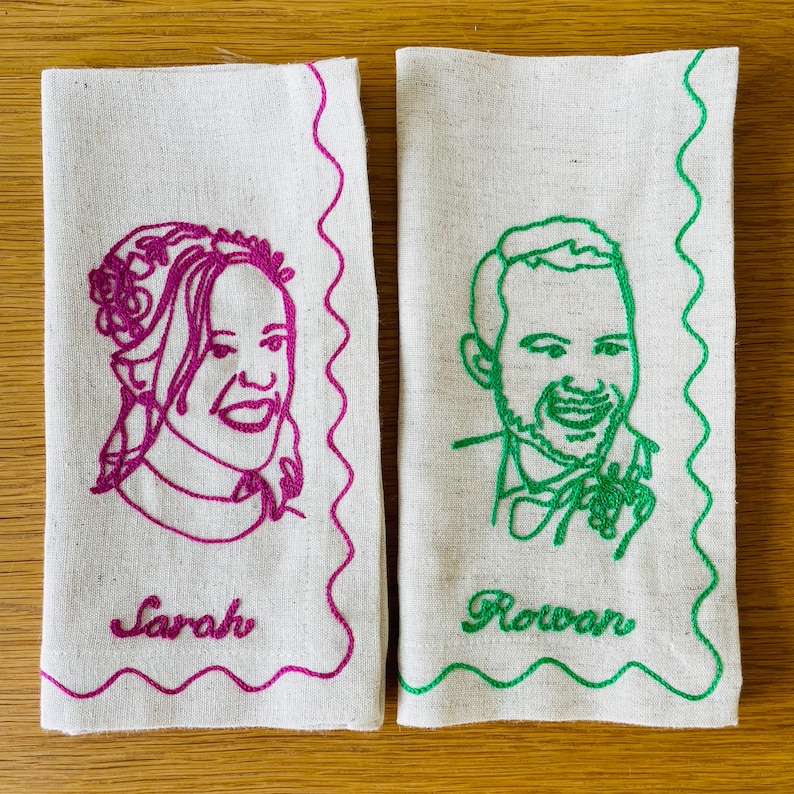 Puede incluir: Dos servilletas de lino blanquecino con retratos bordados. Una presenta un contorno magenta de una mujer con el nombre "Sarah" debajo. La otra tiene un contorno verde de un hombre con el nombre "Rowan". Ambas tienen un borde ondulado decorativo.