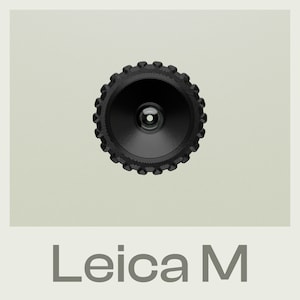 Op de afbeelding: Een zwarte camera lens met een cirkelvormig ontwerp en een kleine, ronde opening in het midden. De tekst "Leica M" is onder de lens afgedrukt.