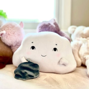May include: A white plush cloud-shaped toy with a smiling face and black eyes. The cloud has a small gray plush pocket on its side.