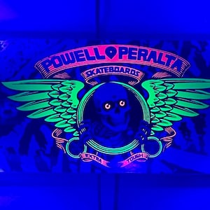 Op de afbeelding: Een zwart en groen skateboardlogo met vleugels, de tekst "Powell Peralta Skateboards" en de woorden "Ride Forever" in een cirkel. Het logo wordt belicht met een blacklight, waardoor het in neonkleuren gloeit.