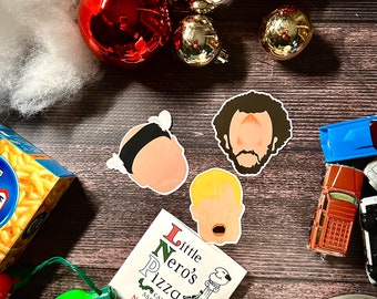 以下が含まれることがあります： ホーム・アローン映画のキャラクターを描いたステッカー3枚。ステッカーは、クリスマスのオーナメントとリトル・ネロのピザの箱が置かれた木製の表面に貼られています。ピザの箱には、「リトル・ネロのピザ」と「コール 555-8885 ノー・フィドリン・アラウンド！」の文字が書かれています。