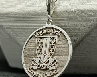 May include: A silver pendant necklace featuring a circular charm with the words "Virginia Tech" and "Corps of Cadets." The charm displays a shield design with a sword and the motto "Ut Prosim." The necklace has a delicate chain.
