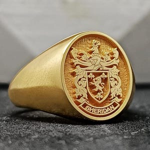 May include: A gold signet ring with an oval face. The face features a detailed crest with a shield, lion, and the name "Sheridan" inscribed. The ring has a polished, smooth finish.