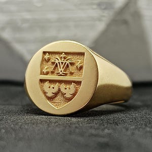 May include: A gold signet ring with a detailed crest. The crest features a central design with two birds below and a floral pattern above. The ring is set against a dark background.