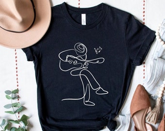 May include: Black t-shirt featuring a white line drawing of a person playing a guitar. The drawing is simple and stylised. The shirt is laid out on a white surface with a hat, jeans, and shoes.