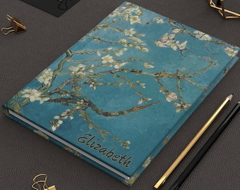 May include: A hardcover journal featuring a blue background with white almond blossom branches and the name Elizabeth printed in dark script. The notebook is accompanied by gold-toned binder clips, a gold pencil, and a black pencil.