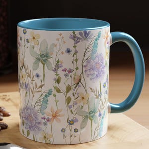 May include: An 11 oz. ceramic mug with a white exterior featuring a watercolor print of various wildflowers in shades of blue, purple, yellow, and green. The mug has a solid blue interior and a matching blue handle, positioned on a wooden surface.