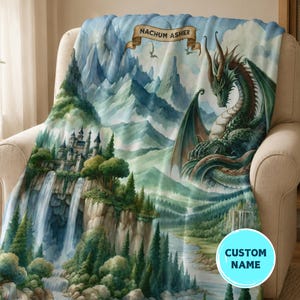 May include: A soft throw blanket featuring a fantasy landscape with a large green dragon, a castle on a cliff with a waterfall, and a banner at the top with the text NACHUM ASHER. The scene includes blue mountains, evergreen trees, and a river.