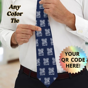 May include: A dark blue tie with a white repeating pattern of QR codes that say "SCAN ME!" on each code. The tie is being held by a person wearing a white shirt and dark blue pants.