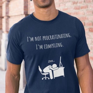 May include: Navy blue t-shirt with white text that reads "I'M NOT PROCRASTINATING, I'M COMPILING." A graphic depicts a person sleeping at a desk with a computer monitor. The shirt is a short-sleeved crew neck.