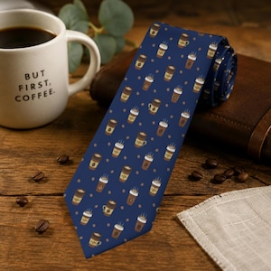May include: A navy blue necktie featuring a repeating pattern of brown and white coffee cups and small coffee beans. The tie is displayed alongside a white ceramic mug with the text BUT FIRST, COFFEE. printed in black on the front surface.