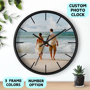 May include: Black round clock with a photo of a couple on a beach. The clock has black hands and black numbers. The text "CUSTOM PHOTO CLOCK" is in the top right corner of the image. The text "3 FRAME COLORS" is in the bottom left corner of the image. The text "NUMBER OPTION" is in the bottom right corner of the image.